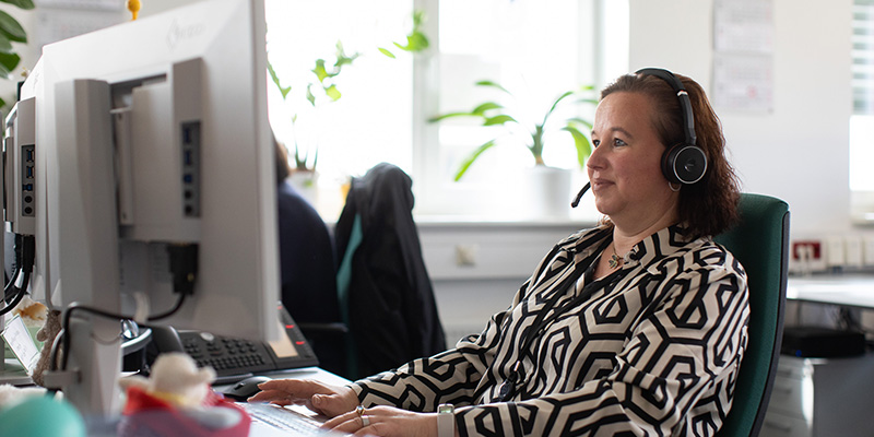Frau in gemusterter Bluse mit Headset sitzt vorm Schreibtisch