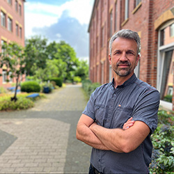 Mann mit grauen Haaren, Bart und kurzaermeligen Hemd, er verschraenkt die Arme